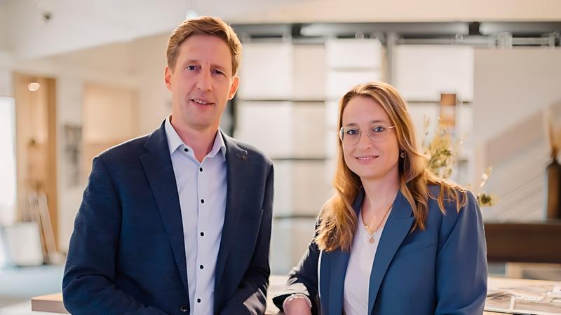 Ein professionelles Business-Team, bestehend aus einem Mann in einem dunkelblauen Sakko und einer Frau in einem passenden Blazer und Jeans, posiert selbstbewusst in einer hellen, modernen Fliesenausstellung.