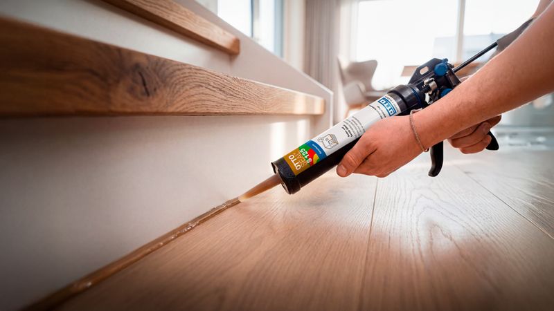 Eine Person trägt mit einer Kartuschenpistole eine Silikonfuge zwischen einer hölzernen Treppenstufe und einem hellen Wandelement auf, um einen sauberen Übergang zu schaffen.