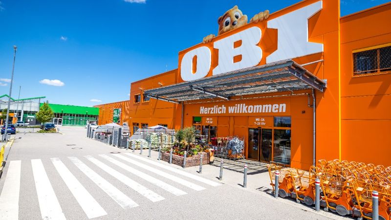 Eine Außenansicht einer OBI-Baumarkt-Filiale bei strahlendem Sonnenschein, mit dem typischen orangefarbenen Gebäude, dem OBI-Logo, dem Biber-Maskottchen und Einkaufswagen im Vordergrund.