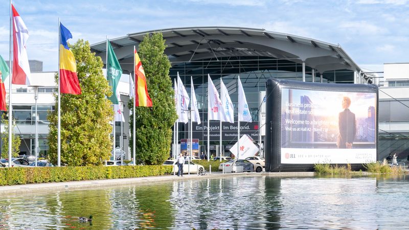 Außenansicht des modernen Messegeländes in München an einem sonnigen Tag. Im Vordergrund befindet sich ein Teich mit Enten, dahinter eine Reihe wehender Fahnen und ein großes, aufblasbares Werbedisplay der Immobilienfirma JLL.