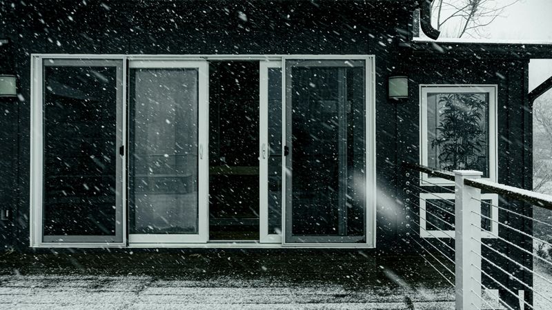 Eine dunkle Hausfassade mit einer modernen Schiebetür auf einer schneebedeckten Terrasse während eines starken Schneefalls.