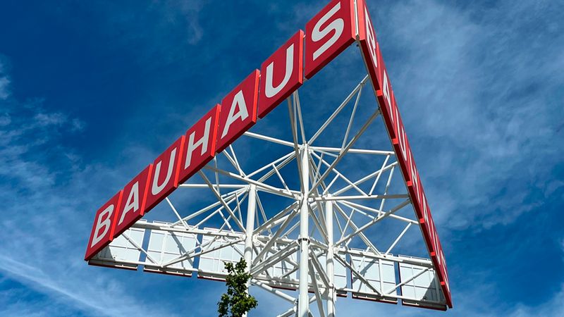 Ein auffälliger, dreieckiger Werbepylon der Baumarktkette Bauhaus, der sich vor einem strahlend blauen Himmel mit leichten Wolken erhebt. Die rote Beschilderung mit weißem Schriftzug wird von einer komplexen weißen Stahlgitterkonstruktion getragen.