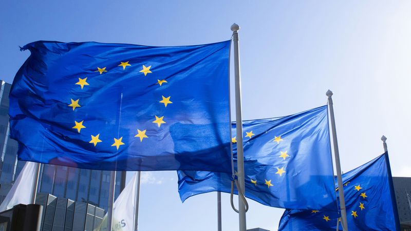 Mehrere blaue Flaggen der Europäischen Union mit dem goldenen Sternenkreis wehen an silbernen Fahnenmasten vor modernen Bürogebäuden bei strahlendem Sonnenschein und blauem Himmel.