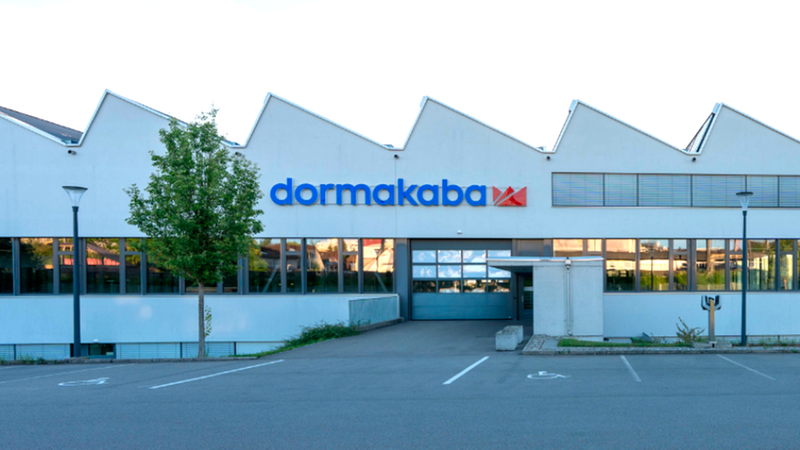 Das Bild zeigt das moderne Firmengebäude von dormakaba mit seiner charakteristischen Sägezahn-Dachkonstruktion und dem Firmenlogo an der Fassade, davor befindet sich ein Parkplatz.