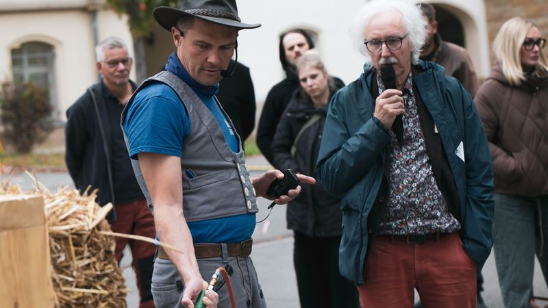 Ein Handwerksmeister mit Cowboyhut erklärt bei einer öffentlichen Veranstaltung in der Stadt eine handwerkliche Tätigkeit, während ein älterer Mann mit weißem Haar und Brille mit einem Mikrofon spricht und eine Gruppe von Zuschauern aufmerksam zuschaut.