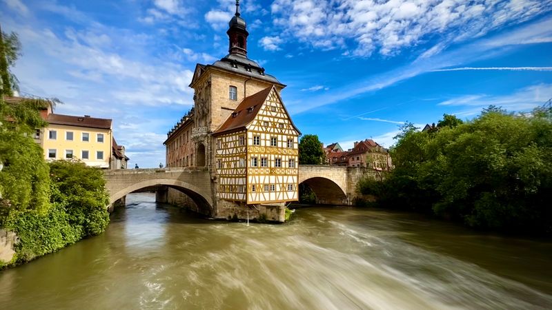 Ein malerischer Blick auf das historische Alte Rathaus in Bamberg, das eindrucksvoll auf einer Brücke mitten in der Regnitz steht, umgeben von grünem Uferbewuchs unter einem strahlend blauen Himmel.