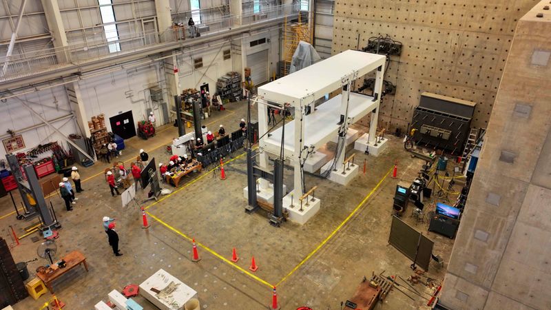 Eine Draufsicht auf eine hochmoderne Versuchshalle, in der ein zweistöckiges Gebäudemodell einem strukturellen Belastungstest unterzogen wird. Ingenieure und Techniker beobachten die Testanordnung, während Sensoren und Messgeräte die Stabilität des Modells überwachen.