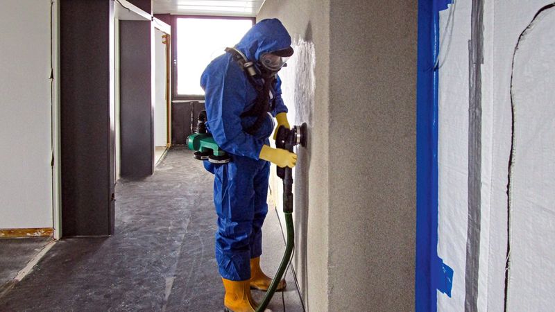 Ein Arbeiter in blauer Schutzbekleidung und mit Atemschutzmaske bearbeitet eine Wand mit einer professionellen Wandschleifmaschine während einer Gebäudesanierung.
