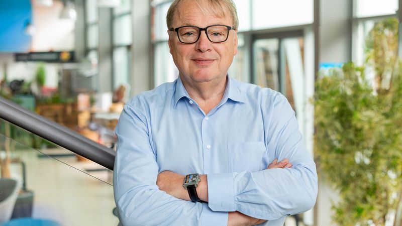 Ein lächelnder Geschäftsmann mit Brille, gekreuzten Armen und blauem Hemd steht in einem Bürogebäude.