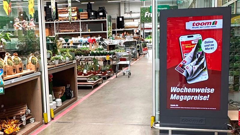 Ein Blick in den Eingangsbereich eines toom Baumarkts, bei dem ein großes, digitales Werbedisplay mit der Aufschrift 'Wochenweise Megapreise!' im Vordergrund steht, während im Hintergrund Pflanzen und Gartenartikel zu sehen sind.