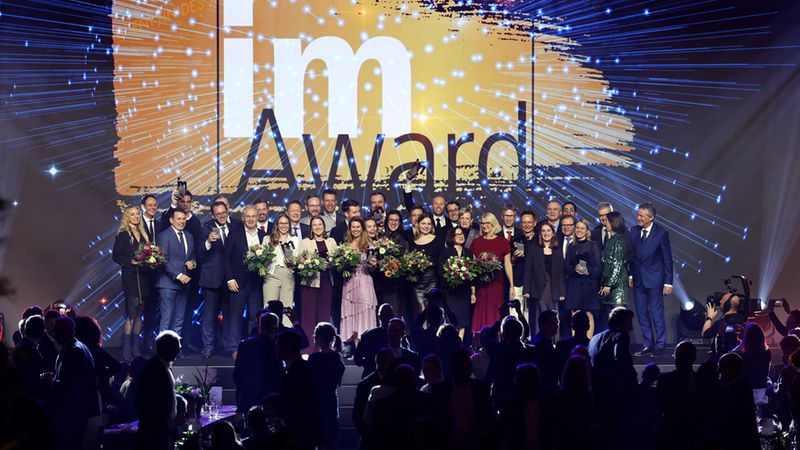 Eine große Gruppe von Preisträgern steht glücklich auf einer Bühne bei der im-Award Preisverleihung vor einer imposanten, digital gestalteten Hintergrundgrafik.