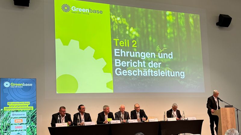 Eine Gruppe von Führungskräften der Firma Greenbase sitzt während der Jahreshauptversammlung an einem langen Tisch auf der Bühne, während ein Redner am Podium den zweiten Teil der Veranstaltung zur Ehrung und dem Bericht der Geschäftsleitung präsentiert.