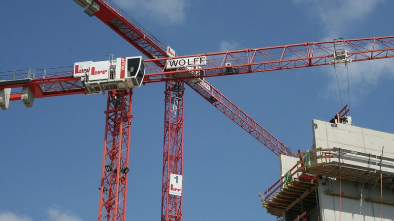 Ein detaillierter Blick auf einen leuchtend roten Wolff-Turmdrehkran auf einer Baustelle vor einem klaren blauen Himmel, daneben ein im Bau befindliches Betongebäude.