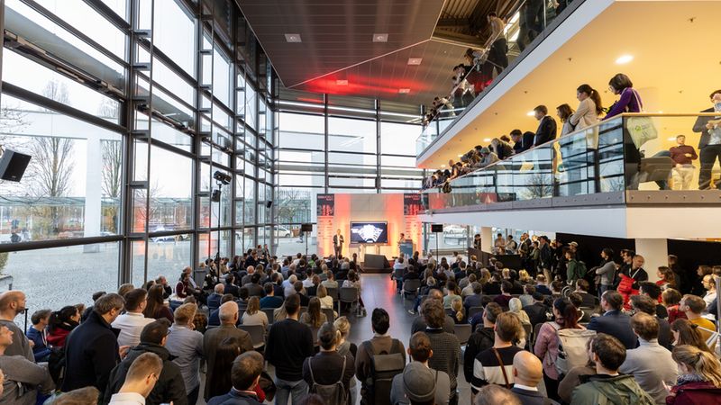 Ein weitläufiger Innenraum mit Glasfassade bei der BIM World Konferenz in München, bei der ein Redner auf einer Bühne vor einem großen Publikum spricht, während weitere Teilnehmer von einer Galerie aus zuschauen.