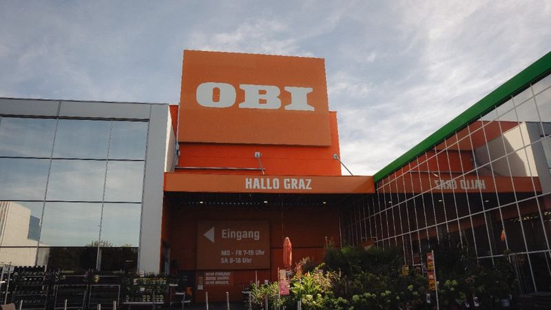 Eine Außenaufnahme der OBI-Baumarkt-Filiale in Graz mit dem markanten orangefarbenen Logo, dem Eingangsbereich und einer Pflanzenabteilung im Vordergrund unter einem leicht bewölkten Himmel.