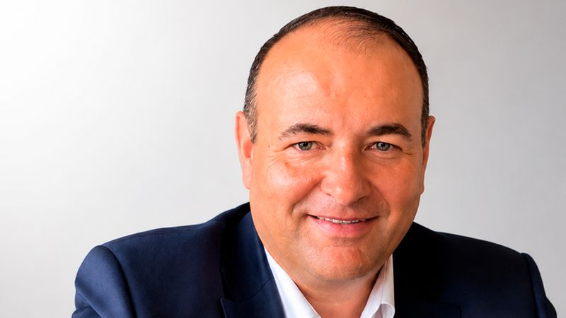 Ein freundlich lächelnder Unternehmer mittleren Alters im blauen Anzug und weißem Hemd, der selbstbewusst an einem Tisch sitzt und in die Kamera blickt.