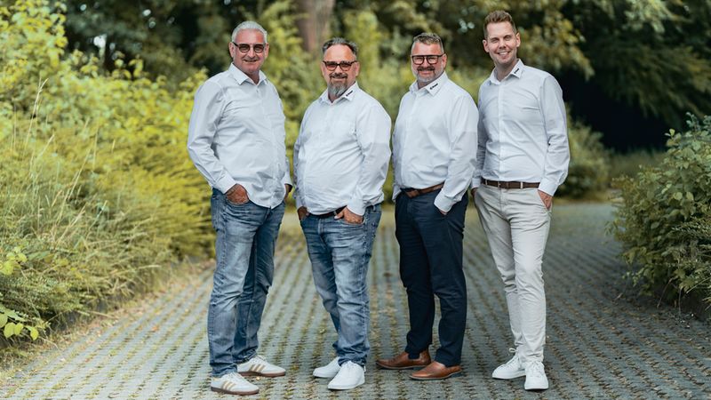 Ein professionelles Foto von vier Männern als Business-Team, die in einer parkähnlichen Umgebung mit grünen Bäumen im Hintergrund stehen. Alle Männer tragen weiße Hemden und lächeln freundlich in die Kamera.