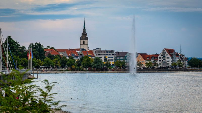 Ein malerischer Blick auf die Stadt Friedrichshafen am Bodensee, bei dem die markante Kirche mit ihrem hohen Turm die Kulisse beherrscht, während im Vordergrund eine hohe Wasserfontäne mitten im See das Stadtbild ergänzt.