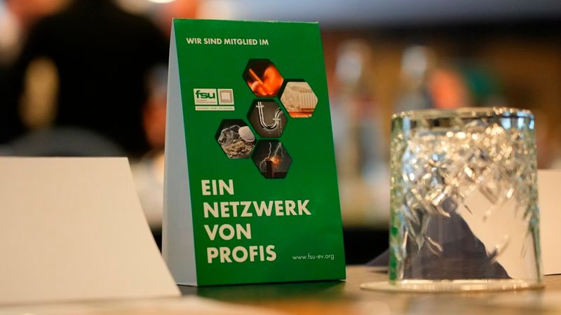 Ein grüner Tischaufsteller mit der Aufschrift 'Wir sind Mitglied im' und 'Ein Netzwerk von Profis' der FSU, platziert auf einem Konferenztisch neben einem Trinkglas.