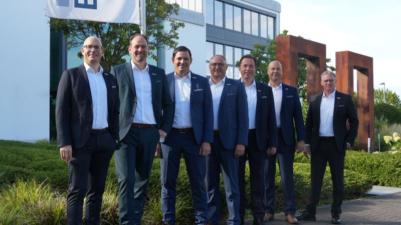 Ein professionelles Management-Team bestehend aus sieben Männern in Anzügen posiert gemeinsam vor einem modernen Bürogebäude mit Firmenflagge.