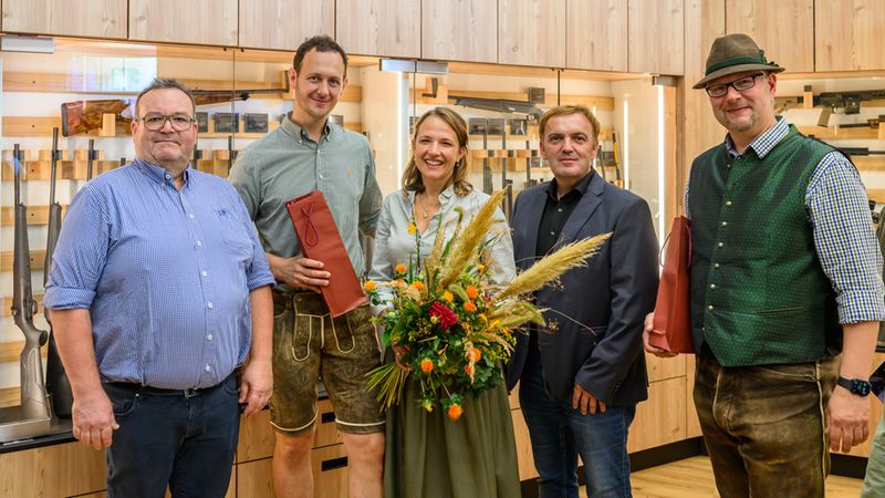 Eine Gruppe von fünf Personen steht lächelnd in einem Waffengeschäft, in dem Gewehre in Vitrinen präsentiert werden. Eine Frau in der Mitte hält einen großen Blumenstrauß, während zwei Männer neben ihr Geschenktüten halten.