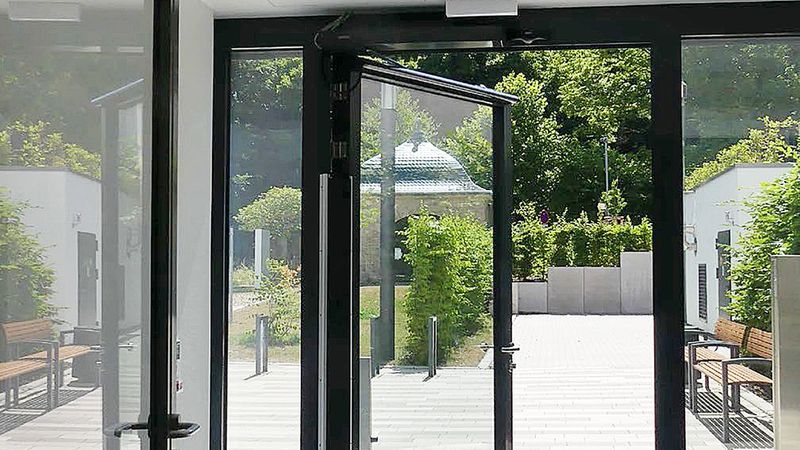 Ein Blick aus dem Inneren eines modernen Gebäudes durch eine geöffnete schwarze Glastür in einen sonnigen Außenbereich mit einer gepflasterten Terrasse, Sitzbänken und einer Parkanlage mit Bäumen im Hintergrund.
