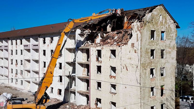 Ein großer Abbruchbagger reißt bei strahlend blauem Himmel das Dach eines mehrstöckigen Wohngebäudes ab.