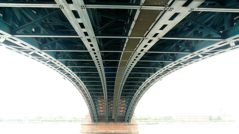 Eine architektonische Perspektive von unten auf eine massive stählerne Bogenbrücke, die über einem Fluss verläuft und den Himmel überdeckt.