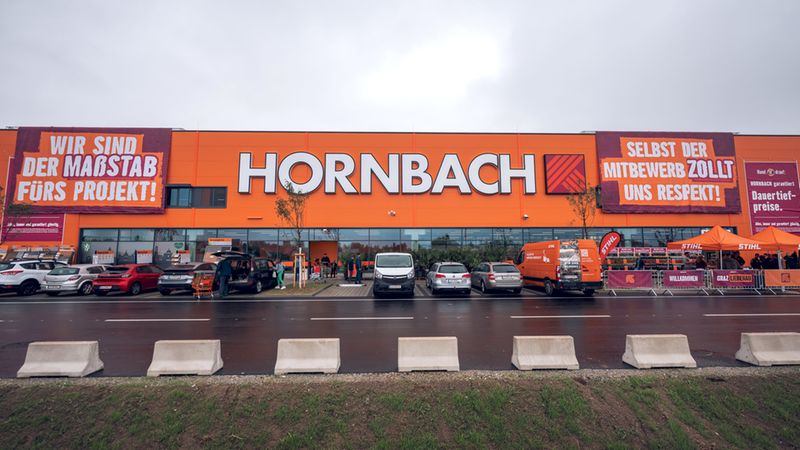 Eine Außenansicht der orangefarbenen Fassade einer Hornbach-Baumarkt-Filiale in Graz-Liebenau mit großen Werbebannern und einem davor liegenden Parkplatz an einem bewölkten Tag.