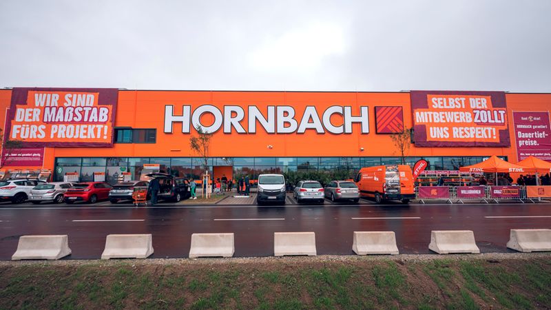 Eine Außenansicht der orangefarbenen Fassade einer Hornbach-Baumarkt-Filiale in Graz-Liebenau mit großen Werbebannern und einem davor liegenden Parkplatz an einem bewölkten Tag.