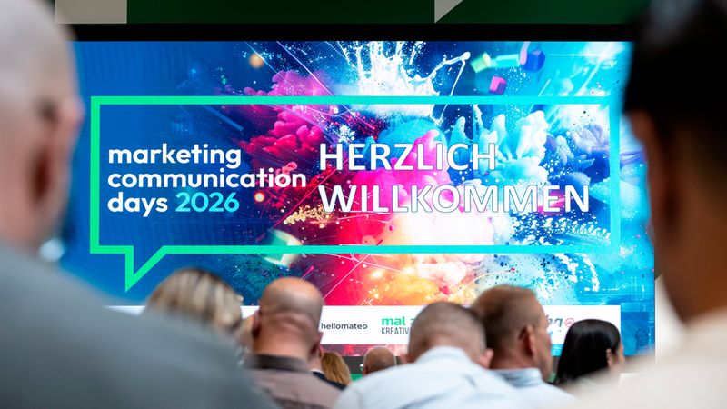 Ein großer Bildschirm auf den Marketing Communication Days 2026 zeigt eine farbenfrohe Begrüßungsgrafik mit der Aufschrift 'Herzlich Willkommen', während das Publikum gespannt auf die Präsentation schaut.
