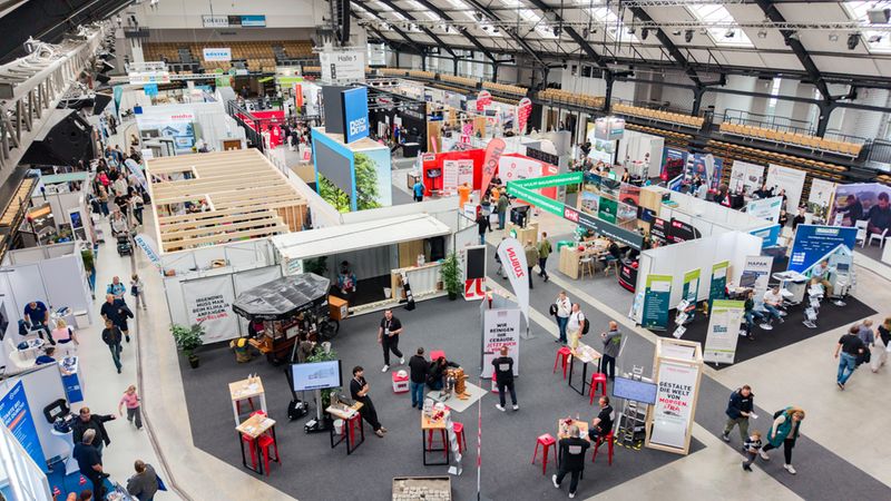 Eine weitläufige Perspektive von oben auf eine belebte Baumesse in einer großen Eventhalle mit zahlreichen Messeständen, Firmenpräsentationen und Besuchern.