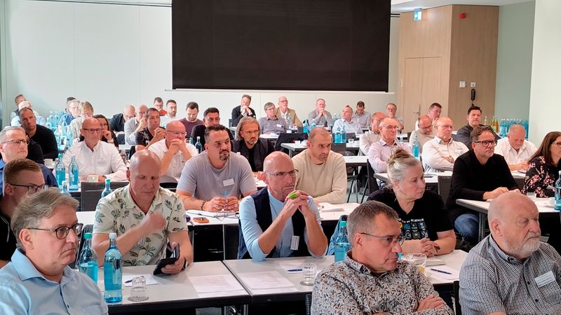 Eine Gruppe von Teilnehmern sitzt in einem modern eingerichteten Schulungsraum an Tischen und hört aufmerksam einem Vortrag zu, während vor ihnen auf den Tischen Wasserflaschen stehen.