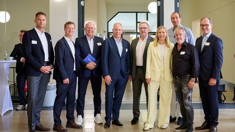 Eine Gruppe von geschäftlich gekleideten Männern und einer Frau posiert für ein gemeinsames Foto in einer hellen, modernen Flur-Architektur bei einem Firmenempfang oder einer Konferenz.