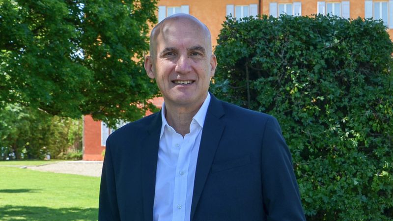 Ein freundlich lächelnder, kahlköpfiger Mann mittleren Alters trägt ein weißes Hemd und ein dunkelblaues Sakko. Er steht vor einem großen, orangefarbenen Gebäude und einem grünen Gebüsch in einem sonnigen Park.