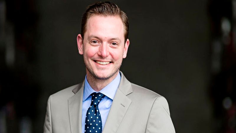 Ein professionelles Portrait eines freundlich lächelnden Geschäftsmannes, der ein hellgraues Sakko mit einem blauen Hemd und einer gepunkteten Krawatte vor einem neutralen, dunklen Hintergrund trägt.