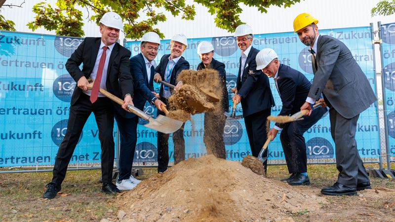 Sechs Geschäftsleute in Anzügen und Schutzhelmen führen gemeinsam einen symbolischen ersten Spatenstich auf einer Baustelle aus. Sie stechen mit Schaufeln in einen Erdhaufen, während im Hintergrund ein blauer Bauzaun mit dem Firmenjubiläums-Logo '100 Jahre IKON' zu sehen ist.