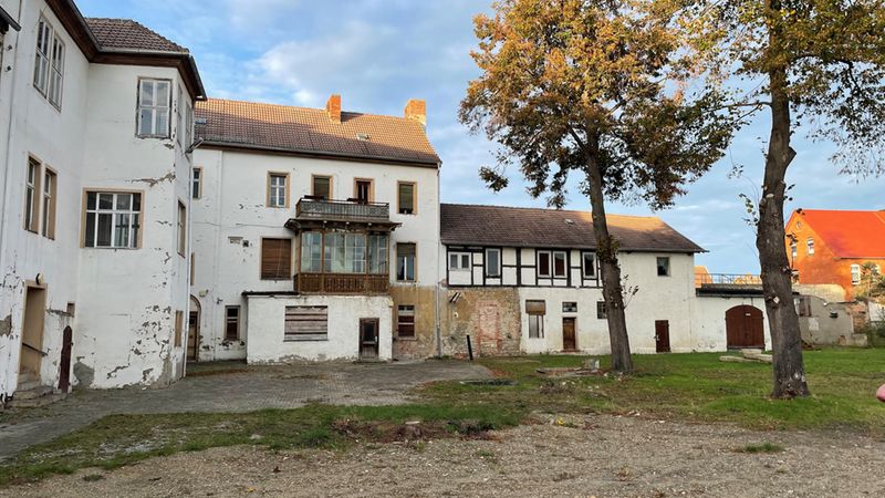 Ein weitläufiger Blick auf die Rückseite eines historischen Herrenhauses mit weiß verputzten Wänden, einem markanten Holzbalkon und anliegenden Fachwerk-Gebäudeteilen, die an einem sonnigen Tag an einen gepflasterten Innenhof grenzen.