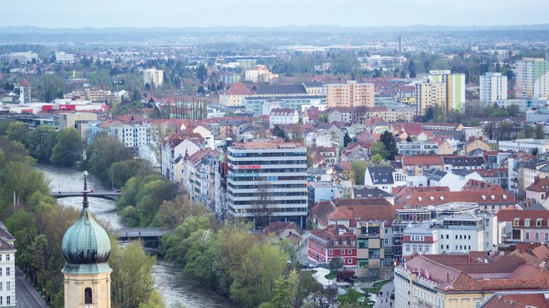 Eine weitläufige Draufsicht auf die österreichische Stadt Graz, die den Fluss Mur, den markanten Turm der Kirche Mariahilf und das dicht bebaute Stadtzentrum mit dem City Tower zeigt.