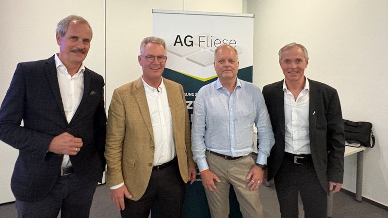 Ein professionelles Foto von vier Männern in Business-Kleidung, die nebeneinander vor einem Banner mit der Aufschrift 'AG Fliese' stehen und lächelnd in die Kamera schauen.