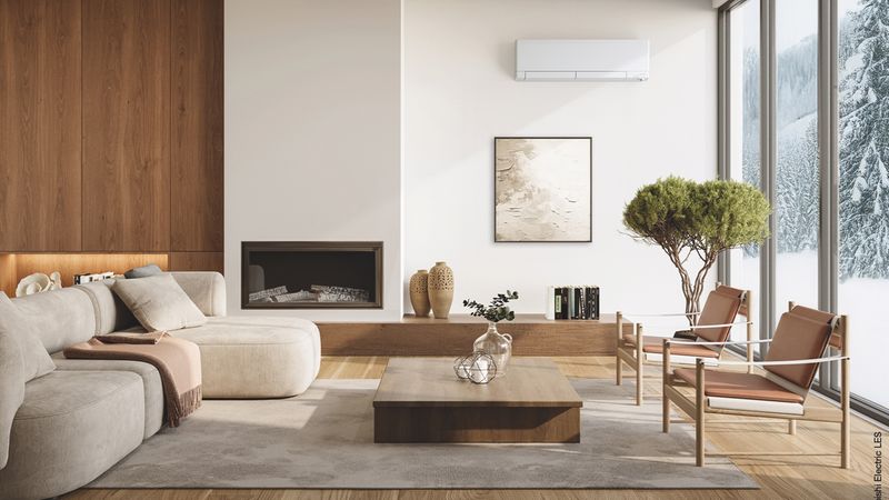 Ein stilvoll eingerichtetes, modernes Wohnzimmer mit einer weißen Luft/Luft-Wärmepumpe, einem gemütlichen Sofa, einem Kamin und großen Fensterfronten, die einen Blick auf eine verschneite Winterlandschaft bieten.