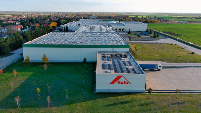 Eine großflächige Luftaufnahme, die den Produktionsstandort und die Lagerhallen der Firma Roto zeigt. Das Industriegebäude ist mit einer weitläufigen Photovoltaikanlage auf dem Dach ausgestattet und liegt eingebettet in eine ländliche Umgebung mit angrenzenden Feldern und einer Ortschaft im Hintergrund.