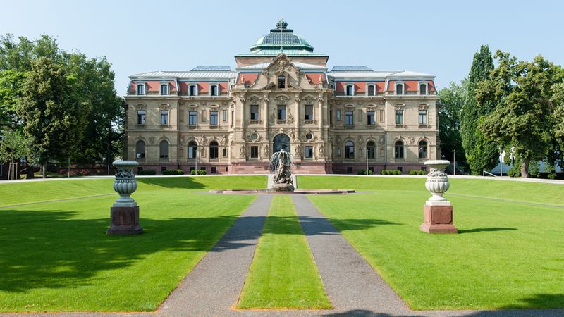 Eine frontale Aufnahme des historischen Justizpalastes in Karlsruhe an einem sonnigen Tag. Im Vordergrund erstreckt sich eine gepflegte Grünfläche mit zwei steinernen Vasen, die einen gepflasterten Weg säumen, welcher direkt auf einen mittig platzierten, künstlerischen Springbrunnen vor dem prächtigen Gebäude führt.