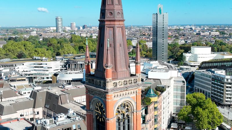 Ein hochauflösender Luftbild-Blick auf den markanten neugotischen Kirchturm der Johanneskirche in Düsseldorf, umgeben von modernen Bürogebäuden, städtischer Architektur und grünen Bäumen unter einem klaren blauen Himmel.