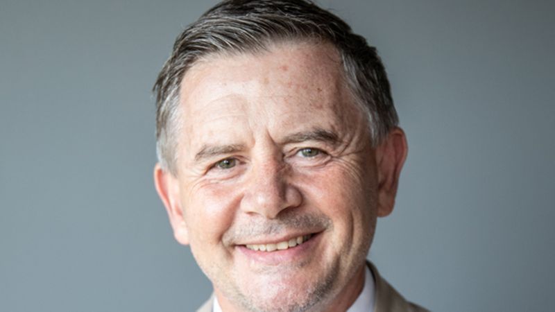 Ein professionelles Nahaufnahme-Portrait eines Mannes mittleren Alters, der freundlich in die Kamera lächelt. Er hat kurzes, graues Haar und trägt ein helles Hemd vor einem neutralen, grauen Hintergrund.
