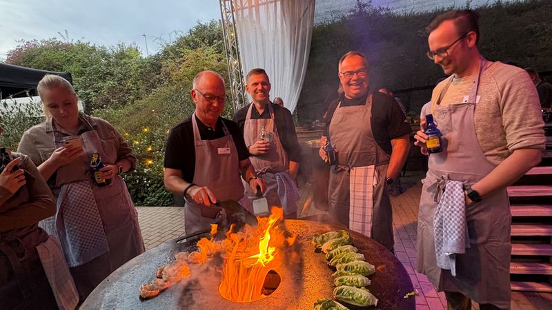 Eine Gruppe von Teilnehmern eines Grillkurses steht um eine Feuerplatte, auf der Salatköpfe gegrillt werden. Die Männer tragen Schürzen und bereiten das Essen über dem offenen Feuer zu, während die Atmosphäre entspannt und gesellig wirkt.