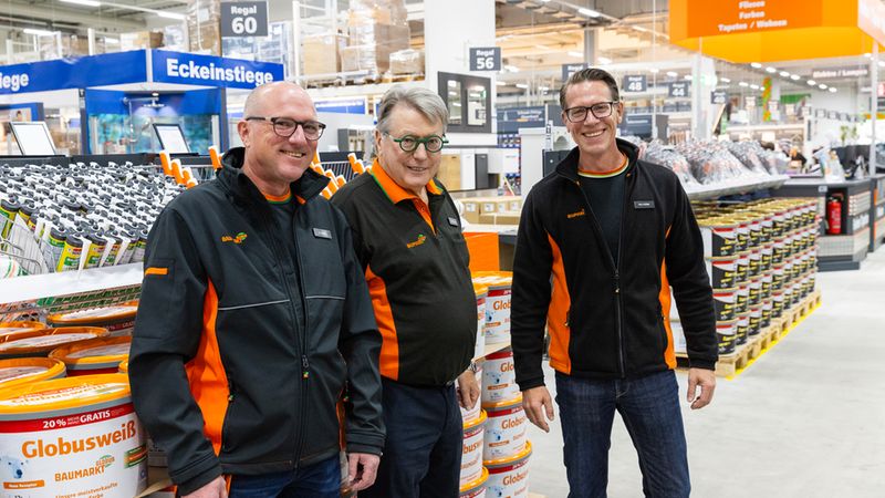 Drei freundliche Mitarbeiter in Arbeitskleidung stehen in einem Globus-Baumarkt vor Regalen mit Farbeimern und Sanitärbedarf.