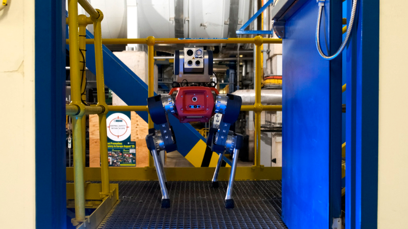 Ein moderner vierbeiniger Industrieroboter mit Sensorkopf steht auf einer metallischen Plattform innerhalb einer industriellen Anlage zur Inspektion bereit.