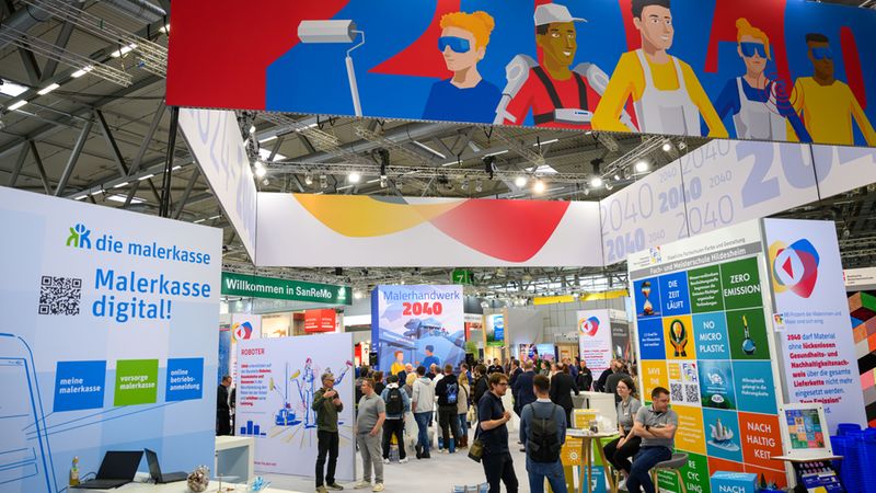 Ein weitläufiger Blick auf einen modernen Messestand auf einer Fachmesse für das Malerhandwerk. Im Zentrum stehen Infotafeln und Aufsteller zum Thema "Malerhandwerk 2040" sowie "die malerkasse", während Besucher und Aussteller in einer lebhaften Messehalle interagieren. Über dem Stand hängt ein großes, buntes Banner mit Illustrationen von Handwerkern.