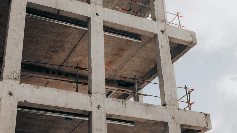 Ein Rohbau eines modernen Beton-Gebäudes, das sich im Bauprozess befindet. Man sieht die tragende Stahlbeton-Skelettstruktur mit mehreren Stockwerken, freiliegende Decken und installierte Gerüste vor einem leicht bewölkten Himmel.